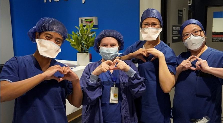 Four nurses in nursing scrubs, holding up their hands in heart shapes and smiling at the camera